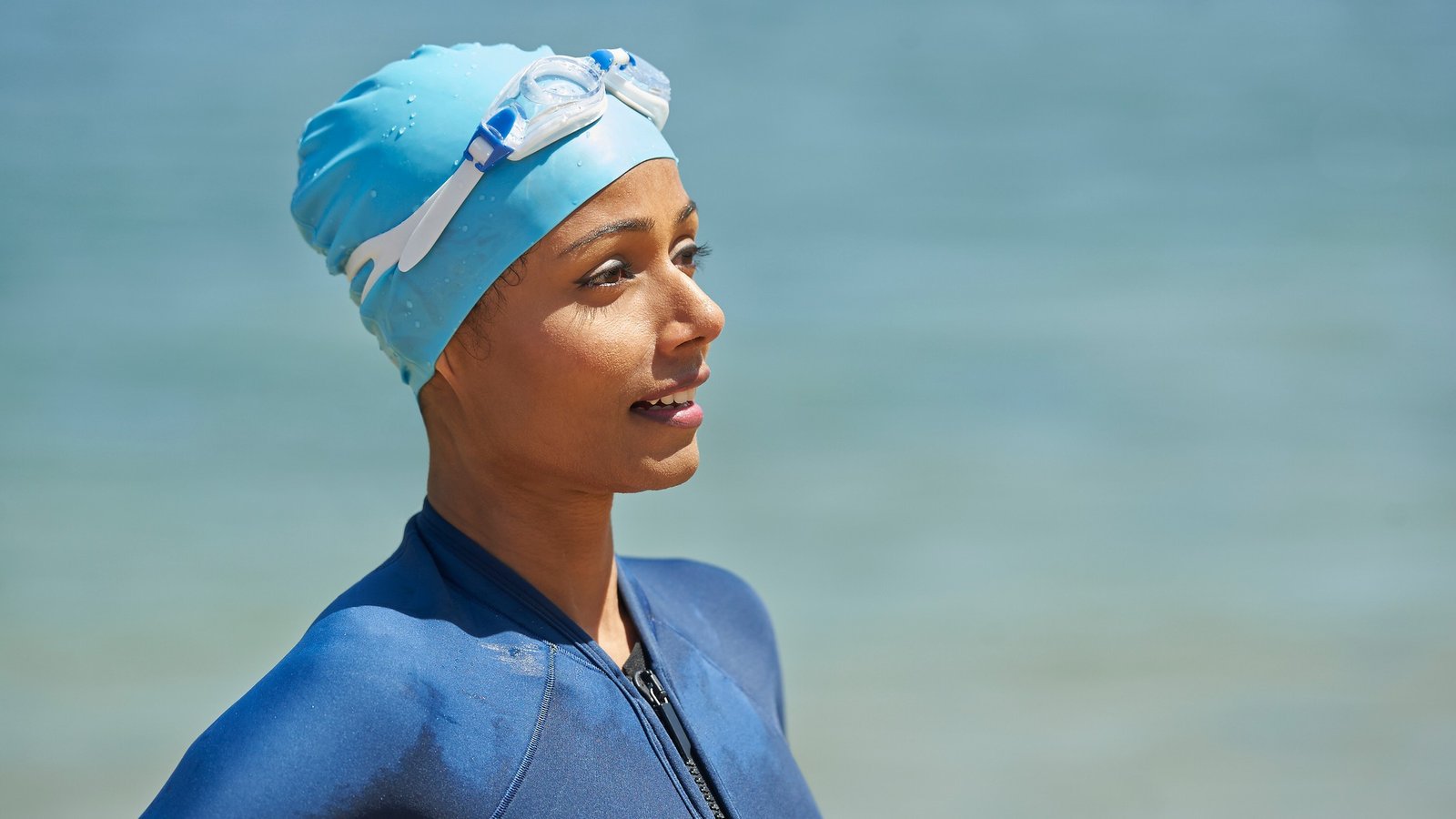 Un nageur se tient devant un plan d'eau naturel.  Elle porte un bonnet de bain sur la tête et elle porte un maillot de bain.  Les deux sont de différentes nuances de bleu.