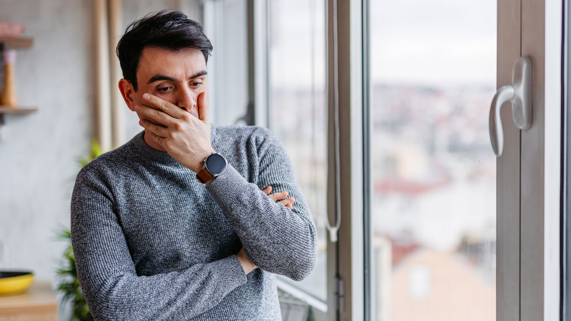 Triste jeune homme debout près de la fenêtre à la maison et contemplant.