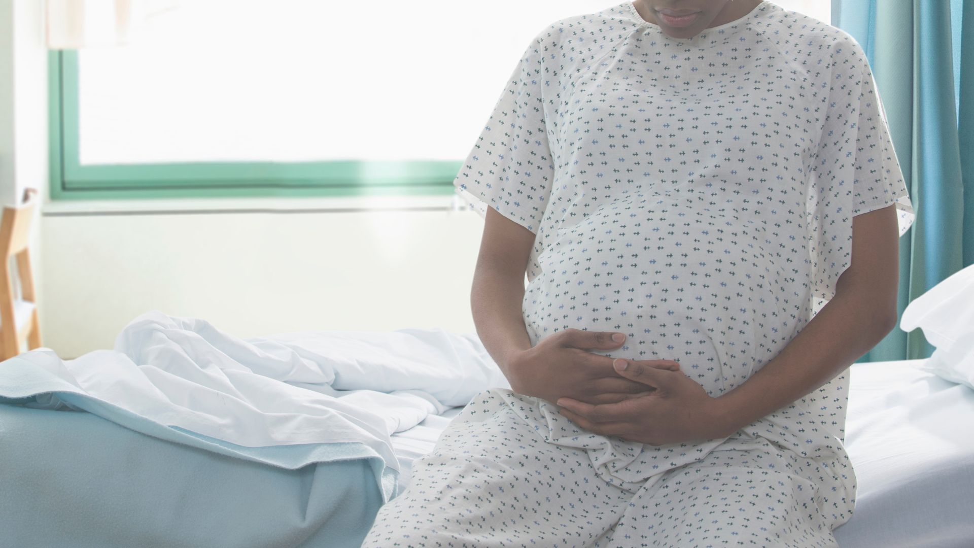 la photo montre une femme enceinte vêtue d'une robe en papier assise sur un lit d'hôpital, les mains sur le ventre