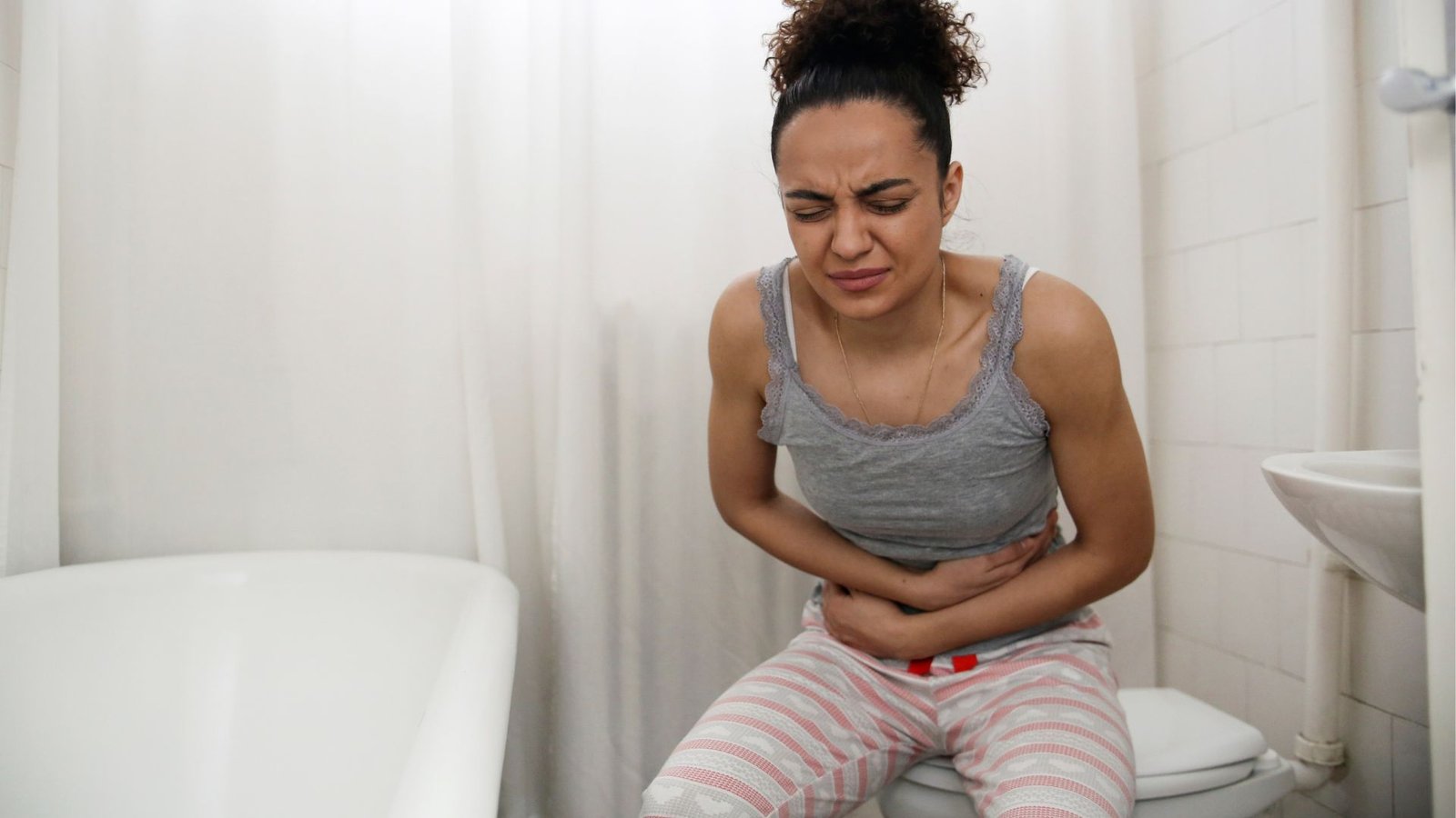 une femme à la peau marron clair est assise dans une salle de bain, serrant son ventre comme si elle souffrait.  Elle porte un débardeur et un bas de pyjama