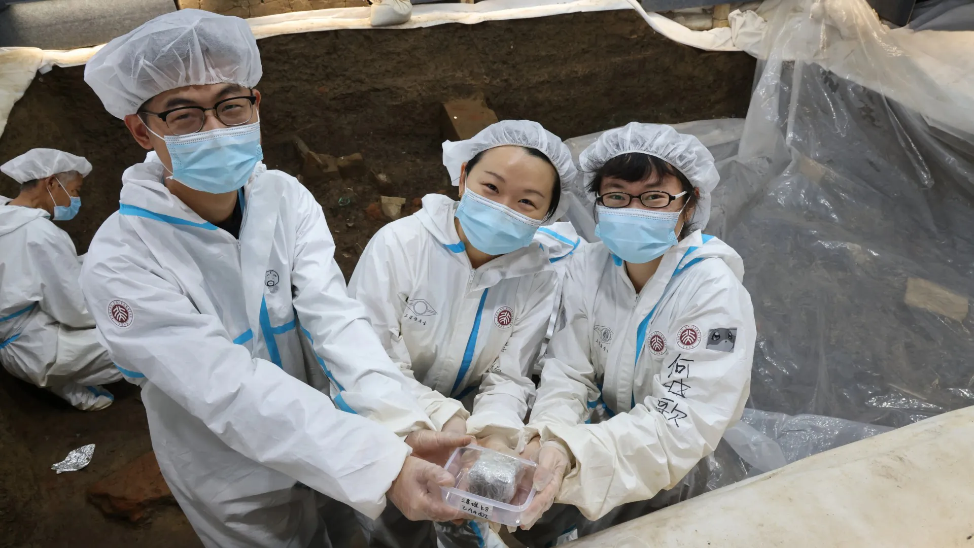 L'archéologue Li Nan (au centre) avec deux membres de l'équipe archéologique de l'Université de Pékin sur le site archéologique de Sanxingdui dans la province chinoise du Sichuan.  (plus une personne de plus travaillant à l'arrière).  Tout le monde porte des couvre-cheveux, des masques faciaux et des combinaisons de protection blanches.