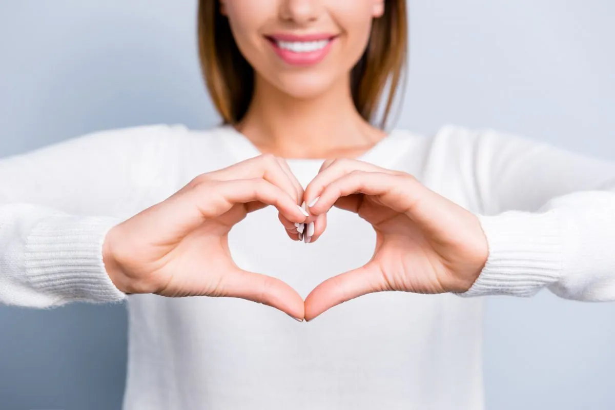 portrait recadré d'une femme souriante en tenue blanche montrant la figure du cœur du symbole d'amour concentré avec les doigts isolés sur fond gris