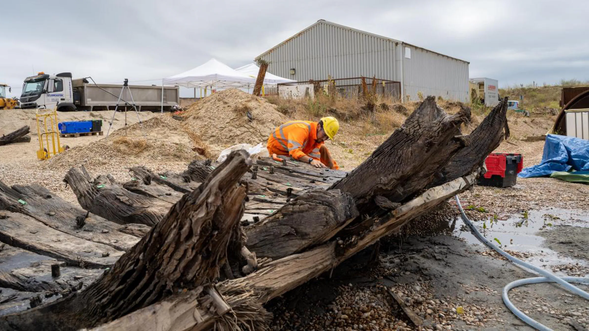 La photo montre une personne vêtue d'une veste orange vif et d'un casque jaune inspectant la coque d'un navire sur la terre ferme